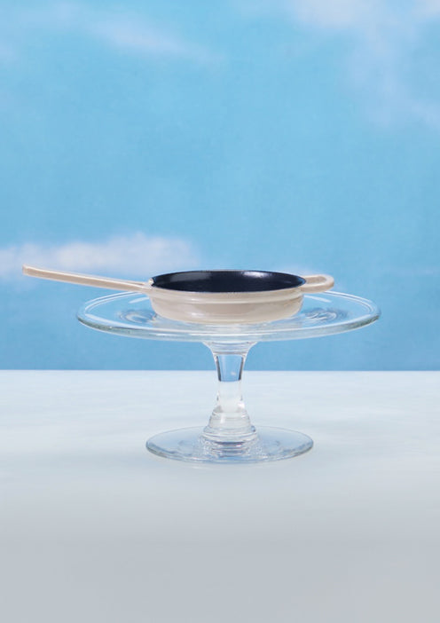 A small black and white frying pan with a handle rests on a clear glass cake stand against a blue sky background.