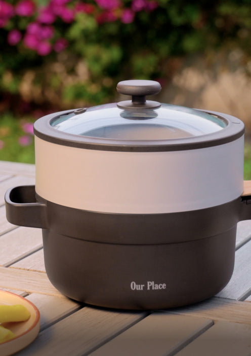 A black and white cooking pot with a glass lid labeled Our Place sits on an outdoor wooden table, with blurred flowers and greenery in the background.
