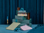 A stack of teal gift boxes with blue bows, a card reading Lets dig in, two amber glasses, a gold pen, a closed notebook, and a folded pink cloth on a blue table.