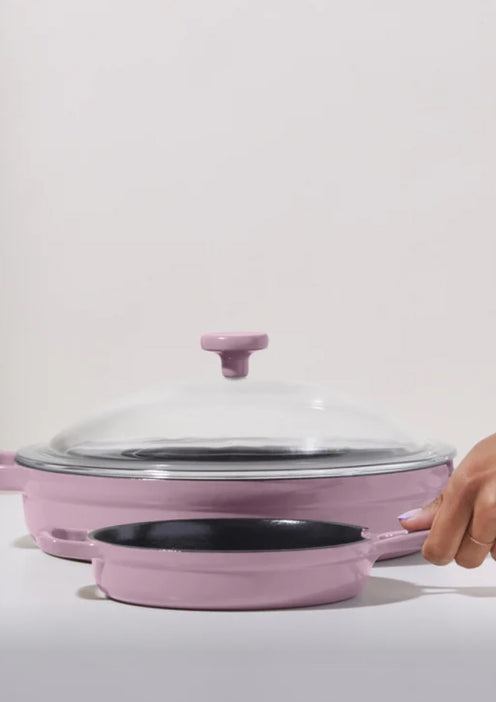 A hand holds the handle of a small pink frying pan stacked in front of a larger pink pan with a glass lid, set against a plain white background.