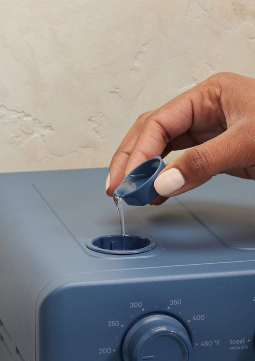 A hand pours liquid from a small cup into the top opening of a blue kitchen appliance with a temperature dial on the front.