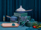 A collection of blue and peach cookware and utensils is arranged on green gift-wrapped boxes, with decorative bows and a single red flower on a blue fabric backdrop.
