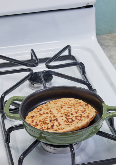 A round flatbread with a cross pattern is cooking in a green cast iron skillet on a white gas stove.