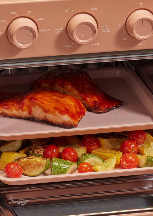 Salmon fillets with sauce and a tray of mixed roasted vegetables are cooking inside a pink oven with visible control dials.