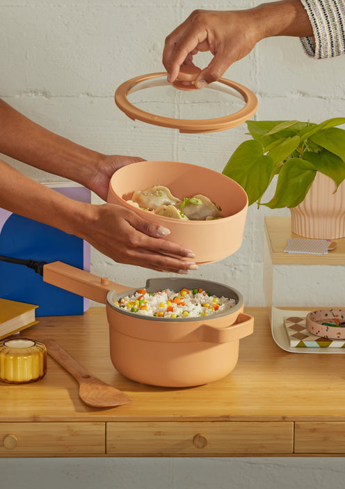 Two hands hold a peach-colored pot and steamer with dumplings; rice with vegetables is in the pot, on a wooden table with books, a plant, and other items.