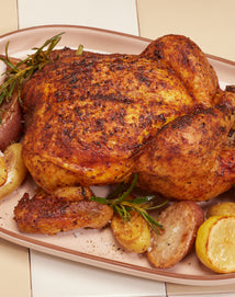 A whole roasted chicken seasoned with herbs, surrounded by roasted potatoes and garnished with sprigs of rosemary on a serving platter.