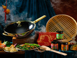 A wok with a glass lid, bamboo steamer, jars of chili sauce, a box with a dragon, plates of food, and utensils are arranged on a table with steam rising.