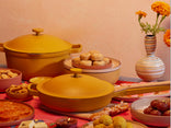 Yellow cookware with lids sits on a table surrounded by various sweets and snacks; a striped vase with marigold flowers is in the background.