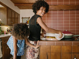 A woman smiles while lifting a lid off a pot in a kitchen, with a child standing beside her, partially bent over the counter.