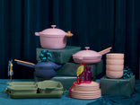 A collection of pastel cookware and tableware, including pots, pans, bowls, and plates, arranged on and around wrapped gift boxes against a dark blue curtain backdrop.