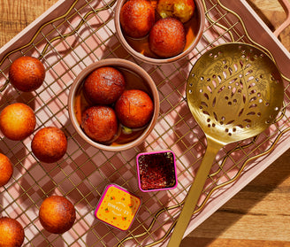 Bowls of gulab jamun on a wire rack with loose gulab jamun, a golden slotted spoon, a tin labeled Diwali ‘23, and a small container of saffron.