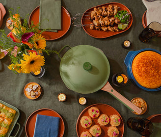 A table set with various dishes, including cornbread, skewers, muffins, a casserole, a green pot, plates, napkins, flowers in a vase, and candles.