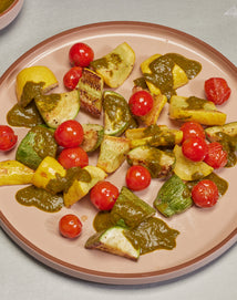 A plate of grilled zucchini, yellow squash, and cherry tomatoes topped with a green sauce, arranged on a light pink plate.