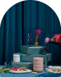 A table with stacked teal gift boxes, blue glassware, pink bowls, a plate with cookies, a small vase with pink flowers, and a hand pouring a drink, set against a blue curtain.