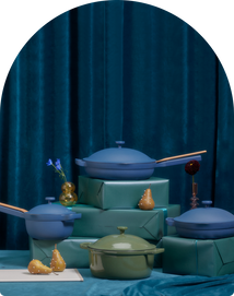 Blue cookware and a green pot are arranged on teal-wrapped boxes and a table, with pears and small vases, set against a blue velvet curtain background.