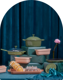 Assorted pink and green cookware and bakeware displayed on teal boxes with a ribbon, a glass cake stand, pastries, and a single pink flower in a vase, against a dark blue curtain backdrop.