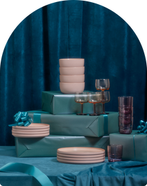 Stacked pink bowls, plates, and assorted glassware arranged on and around teal-wrapped gift boxes, set against a teal fabric backdrop.