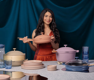 A woman in an orange dress stands behind a table displaying various pastel-colored cookware and tableware, holding a lidded pan and wooden spatula.