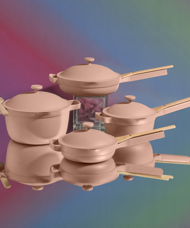 A set of four matte pink pots and pans with wooden handles is arranged on a reflective surface against a multicolored gradient background.
