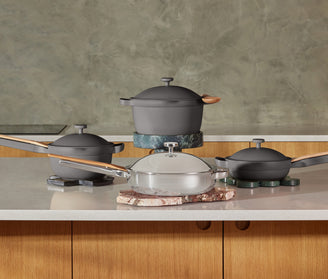 Four modern pots and pans, including gray and stainless steel pieces with wooden handles, are arranged on a kitchen countertop with a green textured wall in the background.