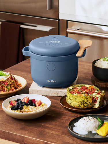 A blue electric cooker sits on a kitchen counter surrounded by five bowls of various rice dishes and a vegetable frittata.