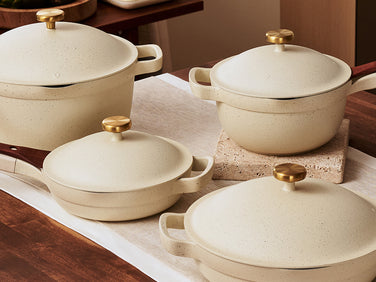 Four cream-colored pots and pans with gold knobs and wooden handles are arranged on a wooden kitchen counter with a white runner and a pot holder.