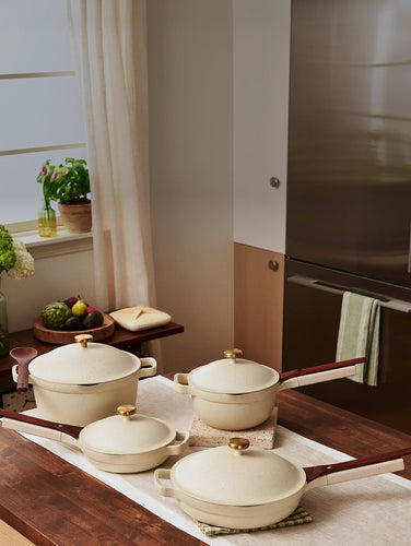 Four cream-colored ceramic pots and pans with wooden handles and gold knobs are arranged on a kitchen counter with other kitchen items in the background.