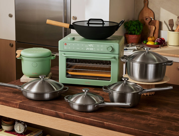 A kitchen counter displays stainless steel pots and pans, a green countertop oven, a green ceramic pot, a black wok, and various kitchen items in the background.