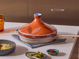A ceramic tagine pot with a patterned lid sits on a textured mat on a kitchen counter, surrounded by small dishes of food and an orange glass.