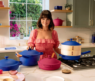 A woman in a pink dress stands in a kitchen behind colorful cookware, including blue and pink pots and pans, with a window showing greenery in the background.