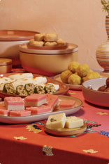 Assorted Indian sweets arranged on plates and bowls atop a red patterned tablecloth, with a vase and stacked containers in the background.
