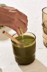 A hand squeezes a lime wedge over a glass of green smoothie with a wooden spoon inside, next to two stacked brown glasses on a light surface.