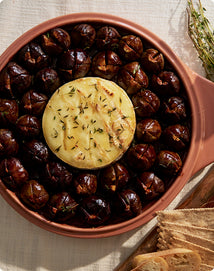 Round terracotta dish with roasted chestnuts and a round piece of cheese on a light fabric background.