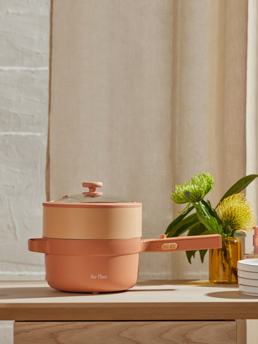 A peach-colored multifunctional pot with a glass lid sits on a wooden counter next to stacked plates, glasses, a yellow vase with flowers, and a yellow candle.