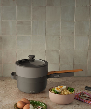 A stacked gray cooking pot with a wooden handle and lid sits on a counter next to a bowl of ramen with egg, greens, and corn. A yellow vase and a plug are also visible.