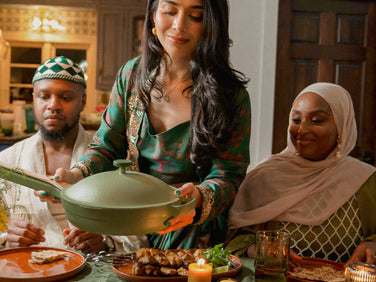 Three people sit at a dining table with plates of food and candles, as a woman in the center serves food from a green pan.