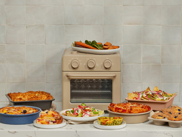 A beige countertop oven is surrounded by various baked dishes, including quiche, lasagna, salad, nachos, muffins, and roasted vegetables against a tiled wall.