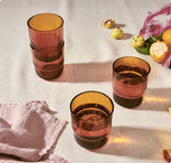 Four amber-colored drinking glasses, two stacked and two with liquid, sit on a light tablecloth near a pink towel, citrus fruits, and kitchen scraps.
