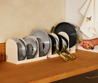 A set of white kitchen organizer stands holding glass lids and cookware handles on a wooden countertop, with a hand holding one of the handles. A beige wall and a white towel with a lemon motif are in the background.