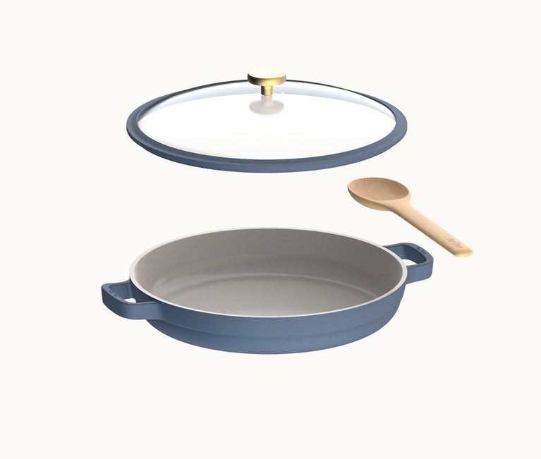Blue round baking dish with two handles, a clear glass lid with a gold knob, and a wooden spoon, all on a plain white background.