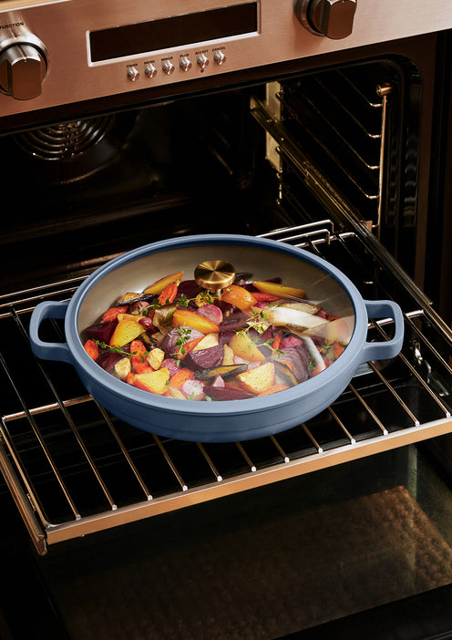 A round casserole dish filled with assorted roasted vegetables is placed on an oven rack, with the oven door open.
