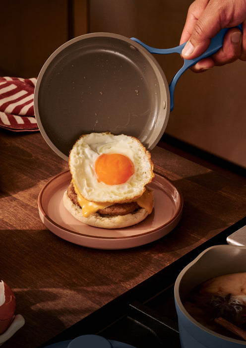 A hand places a fried egg from a pan onto a breakfast sandwich with cheese and sausage on an English muffin, on a wooden countertop.