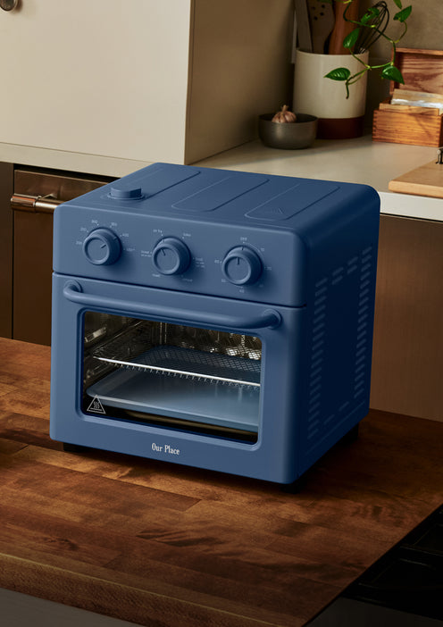 A blue countertop toaster oven with three control knobs sits on a wooden kitchen counter, surrounded by neutral cabinetry and a few plants in the background.