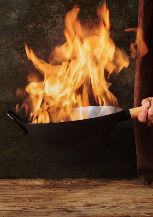 Person holding a black frying pan over an open flame with a dark background