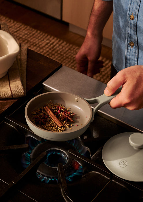 A person holds a small pan with whole spices, including cinnamon sticks and star anise, over a lit gas burner on a stove.
