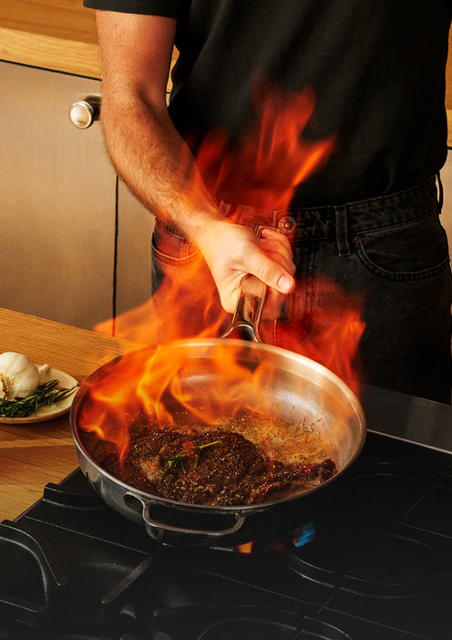 A person in a black shirt and jeans holds a stainless steel pan over a gas stove, with bright orange flames rising from searing food inside the pan in a kitchen setting.