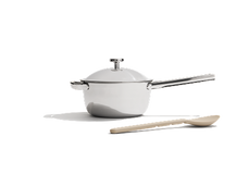 A white saucepan with a lid and long handle is placed next to a beige spoon on a white background.