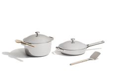 Two stainless steel cookware items with lids, a saucepan and a frying pan, are shown with a wooden spoon and a metal spatula on a white background.
