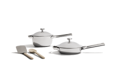 Two stainless steel pans with lids are shown alongside a metal spatula and a wooden spoon, all set against a plain white background.