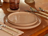 Two stacked beige plates, cream-handled cutlery, a pink glass, and a napkin are arranged on a wooden table.